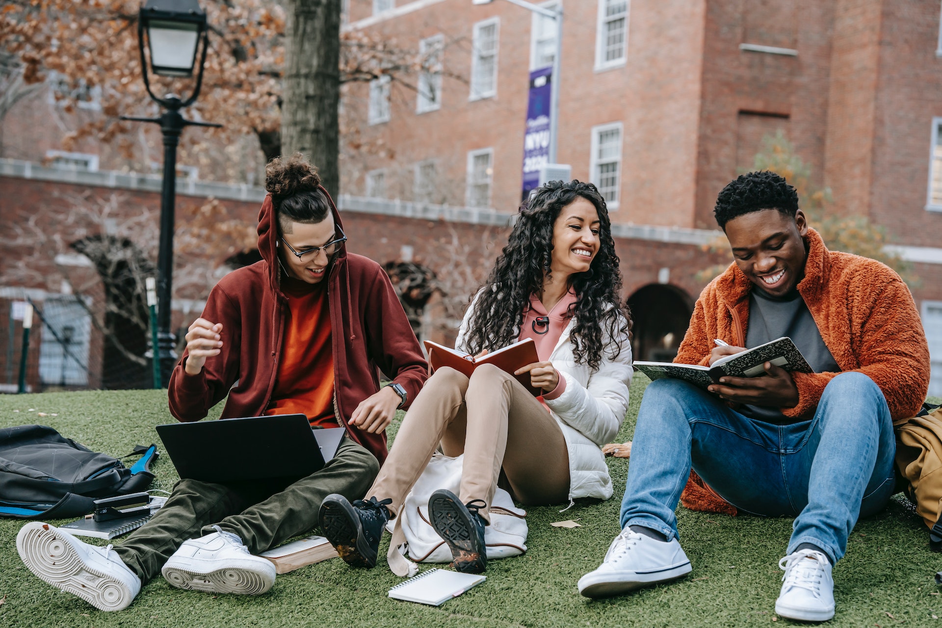 Three classmates studying outdoors on campus with books and laptops