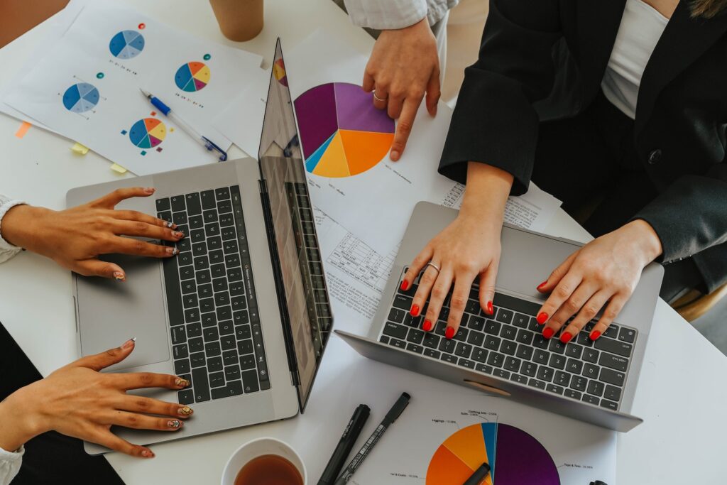 Two women typing on laptops with pie chart graphs on a table.