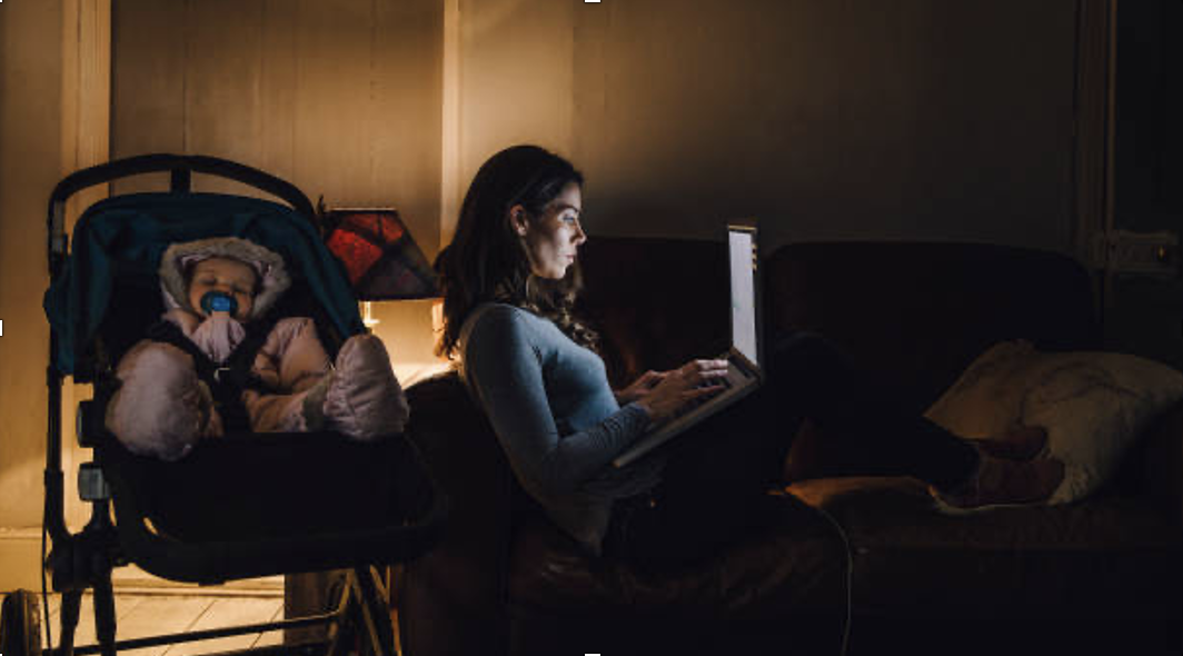 A woman studying on her computer in the dark while a baby sleeps behind her.