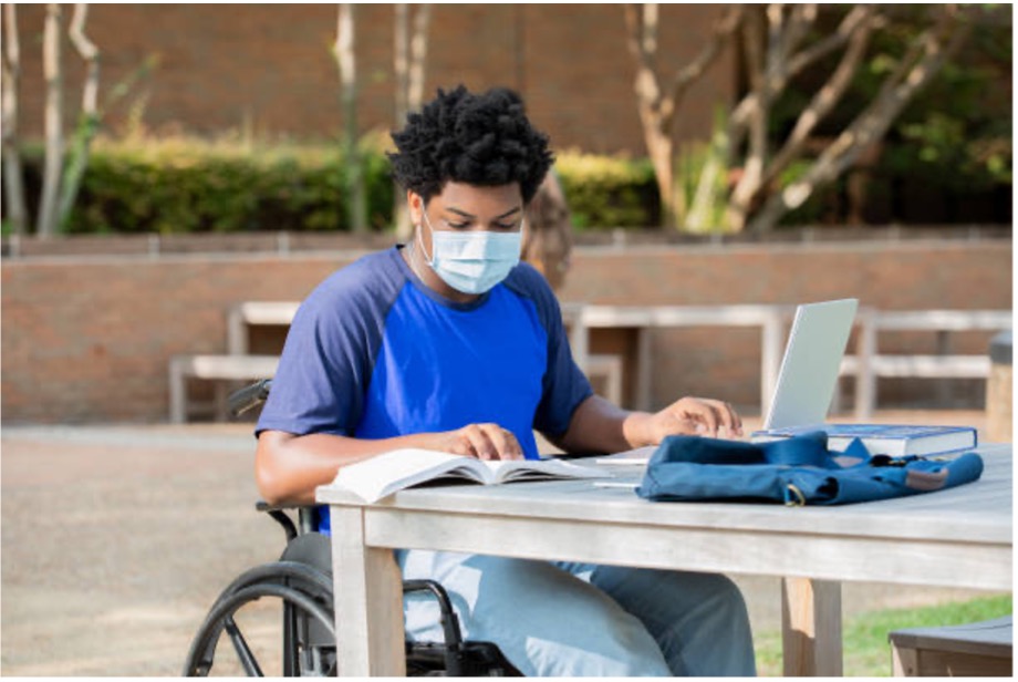 Person wearing a mask sitting at a table reading a book taking notes on a computer.