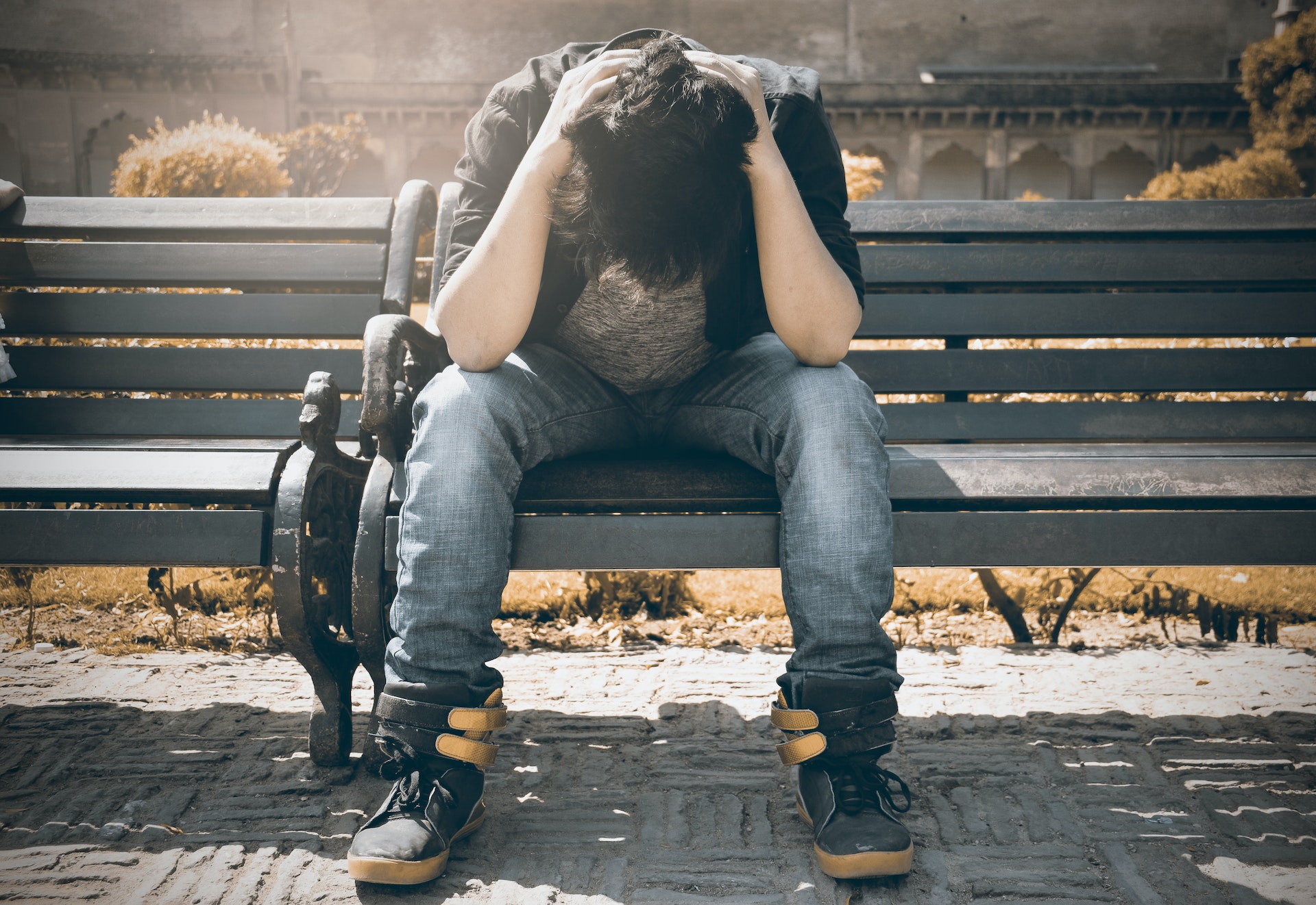 Young man sitting along with his head down on a bench, looking lonely or depressed.