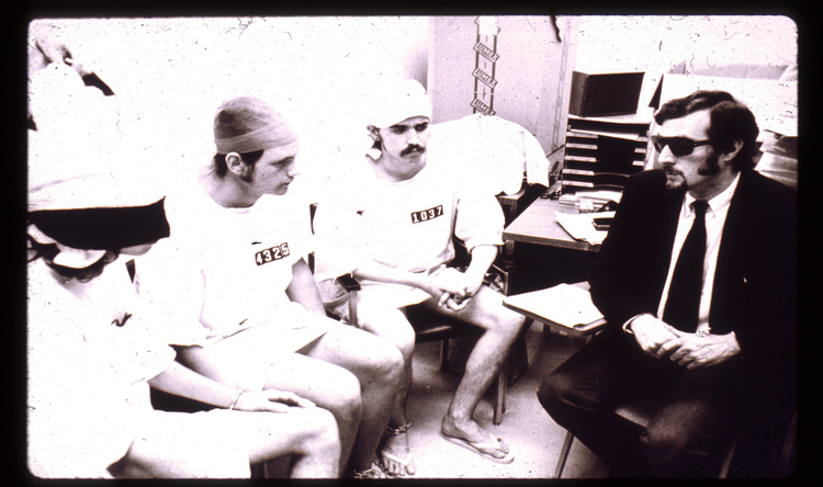 Photograph showing 3 White male college students in prison uniforms as part of the Stanford Prison Experiment. They are seated next to Dr. Zimbardo, who is acting as the superintendent.
