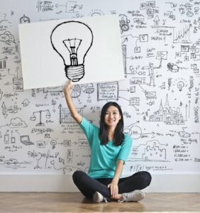 woman sitting on the floor holding a sign with a drawing of a light bulb in front of a wall covered with drawings and diagrams