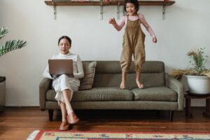 mother sitting on a sofa working on a laptop while daughter jumps on the sofa next to her
