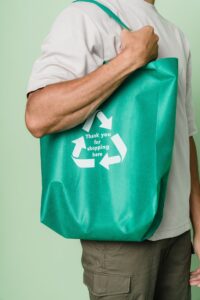person with a reusable shopping tote on their shoulder. The tote has the recycling symbol of arrows arranged in a triangle and say Thank you for shopping here.