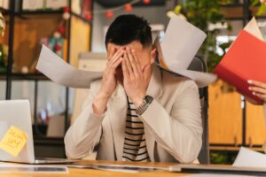 man with his head in his hands, sitting at desk at work with papers being thrown at him