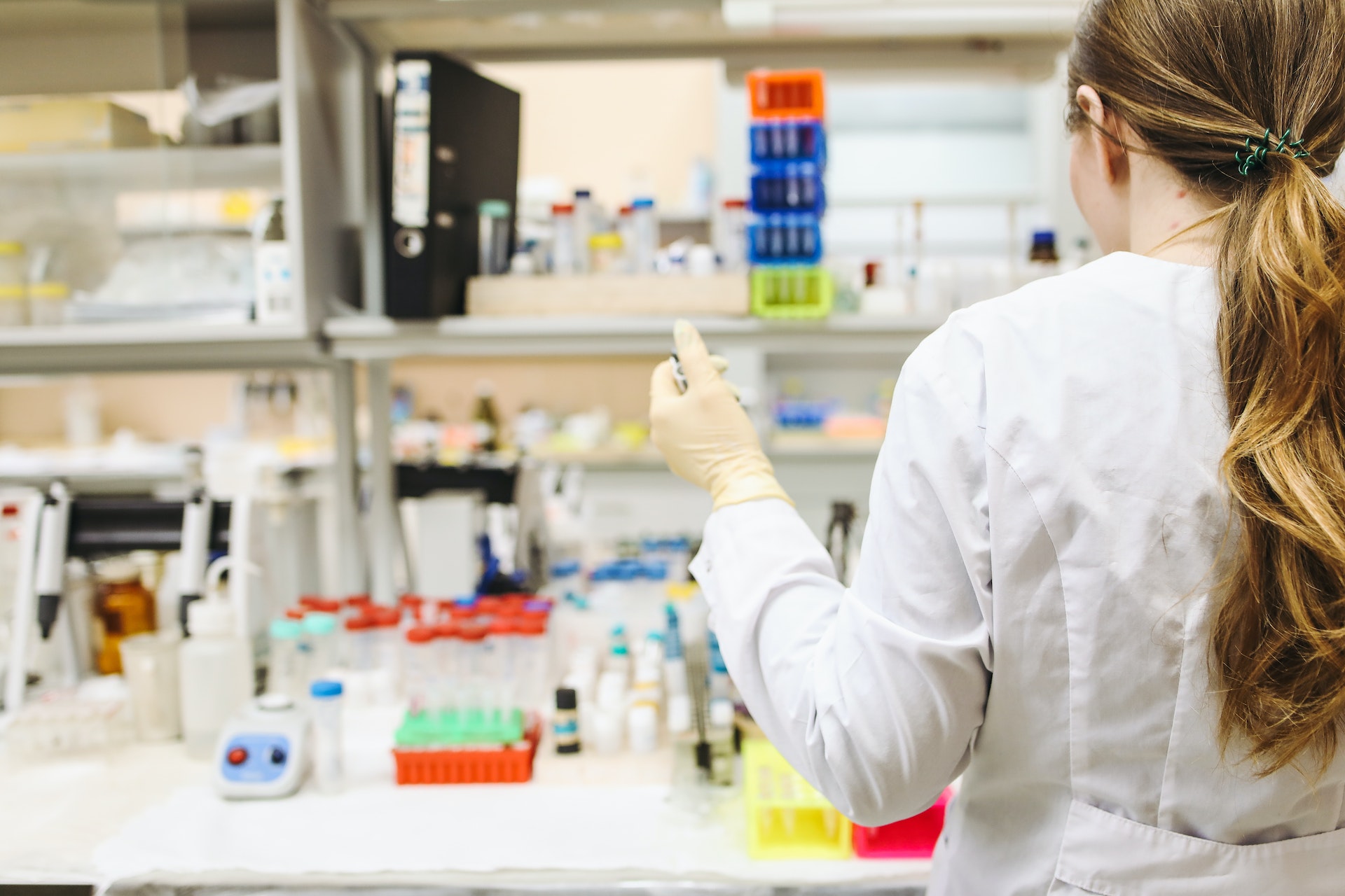 A scientist in a lab coat holds a micropipette facing a bench filled with lab equipment.