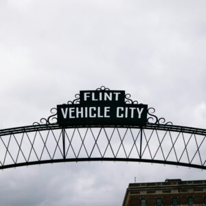 An overhead archway sign reading 'FLINT VEHICLE CITY' against a cloudy sky, with the silhouette of a building in the background.