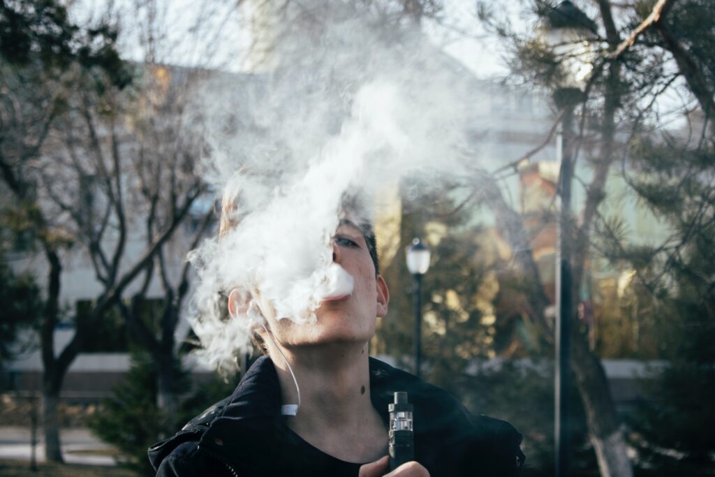 Young man holding a vape pen and blowing smoke in front of his face.