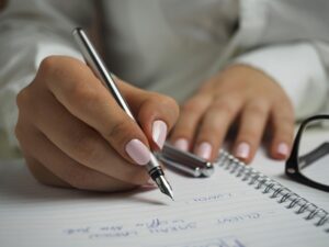 GPT A close-up photo of a person's hands as they write in a spiral notebook with a ballpoint pen.