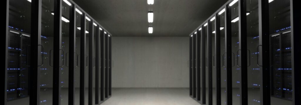 A modern data center corridor with rows of sleek, black server racks on both sides. The server racks are enclosed in glass doors with visible blue indicator lights, highlighting the servers inside.